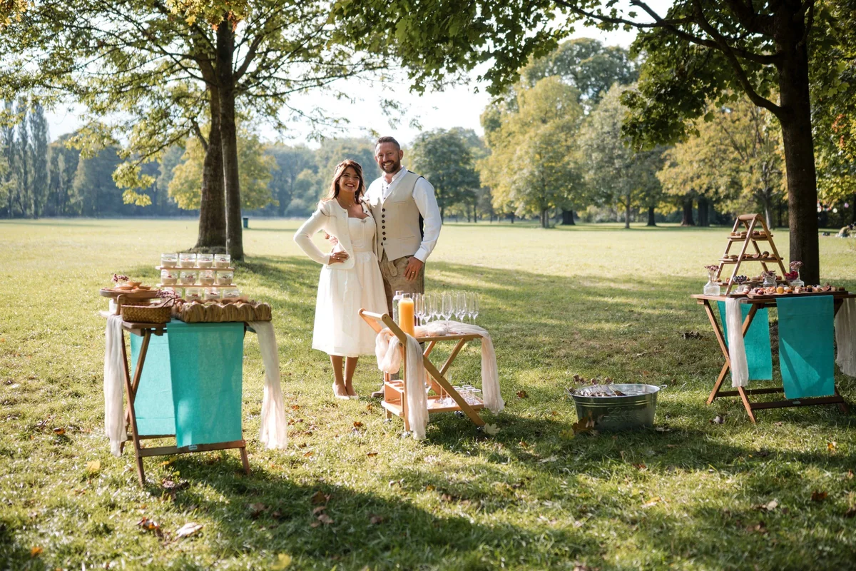 Das Paar in Tracht steht auf einer Wiese im Englischen Garten zwischen Tischen mit Fingerfood.