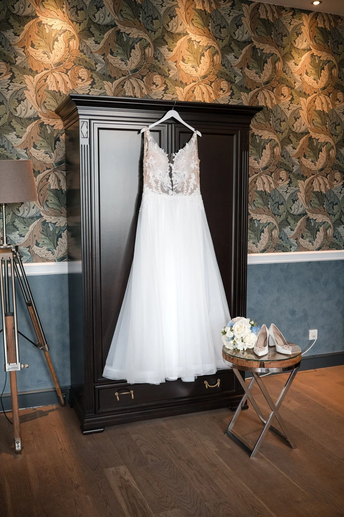 Das Brautkleid hängt an einem dunklen Schrank vor gemusterter Tapete.