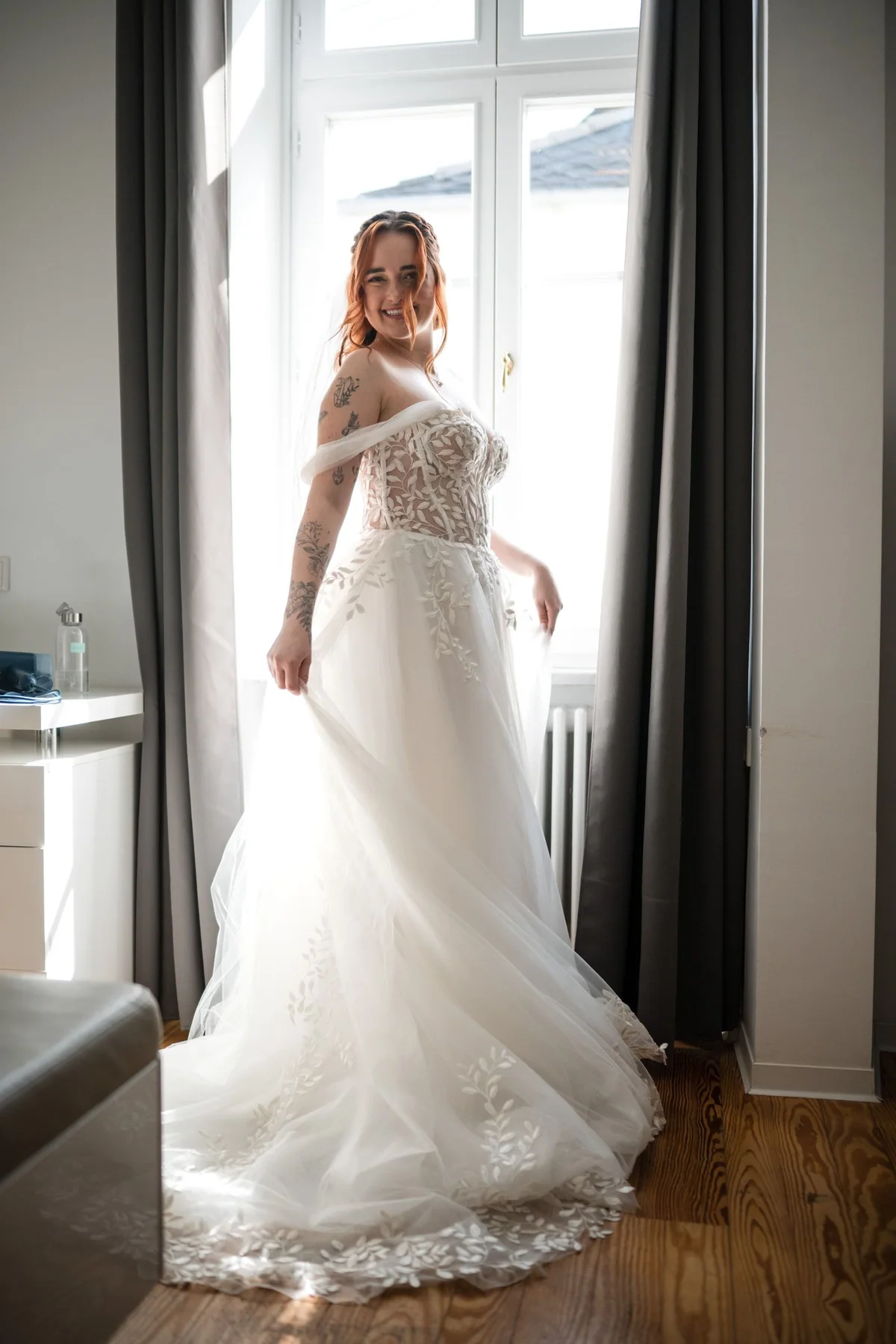 Die Braut steht im Kleid mit Schleppe am hellen Fenster und schaut über die Schulter.
