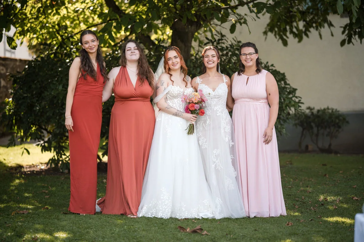 Das Brautpaar mit drei Freundinnen in rostroten und rosa Kleidern im Garten.