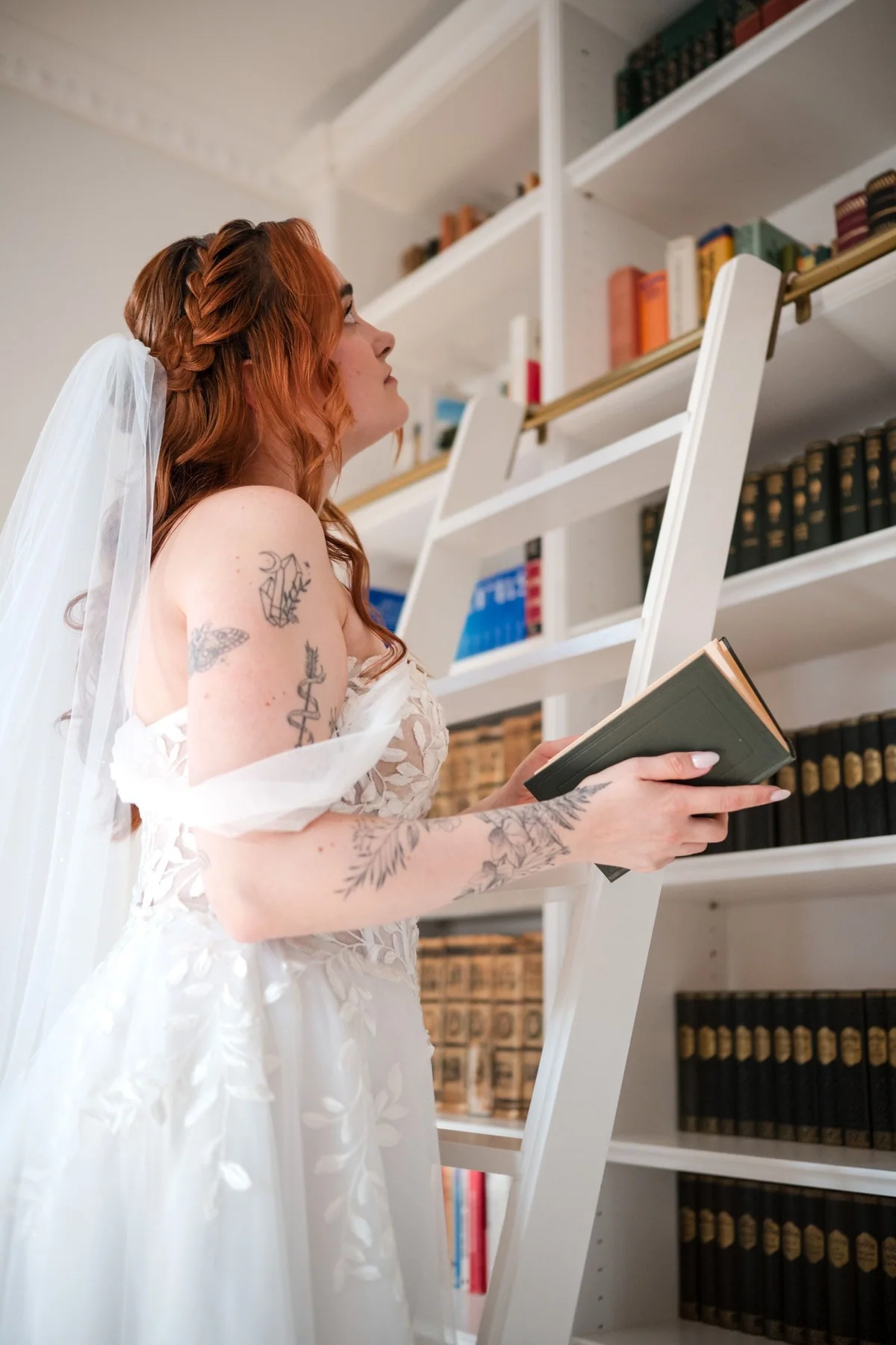 Eine Braut hält am weißen Bücherregal ein altes Buch.