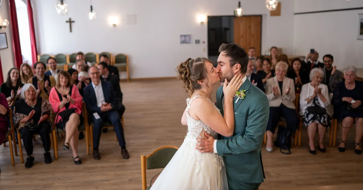 The couple kiss in a bright ceremony room in Bavaria, with guests seated in the background.