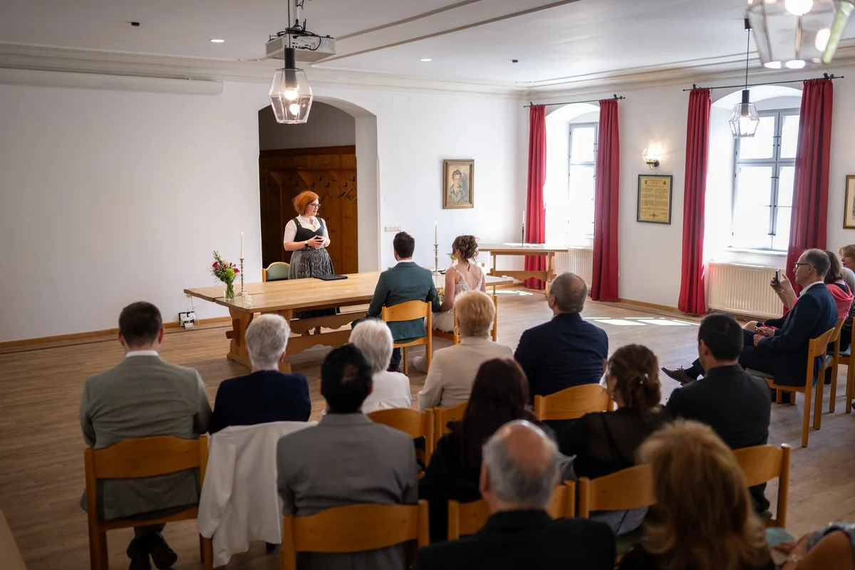 A ceremony in a large, bright room in Bavaria with tall windows and guests behind the couple.