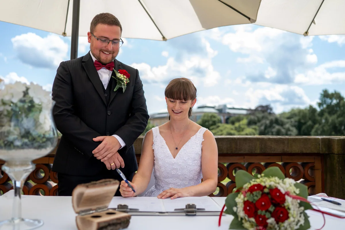 The bride signs under a parasol on the balcony in the botanical garden, the groom laughing beside her.