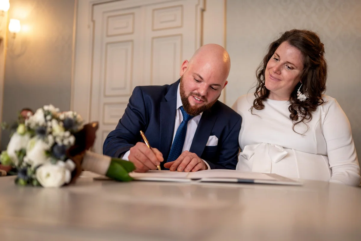 The groom signs at Haus Solms while the bride watches him with a smile, the bouquet lying in front.