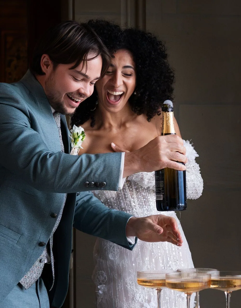 A couple laugh while opening a bottle of sparkling wine over filled glasses.