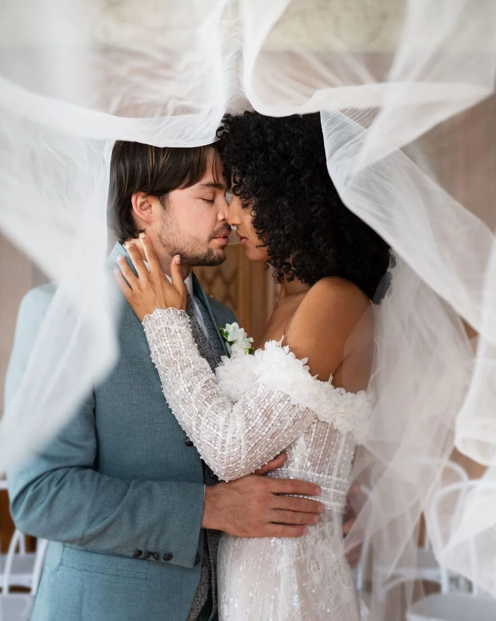 A couple stand forehead to forehead with a veil blowing around them.