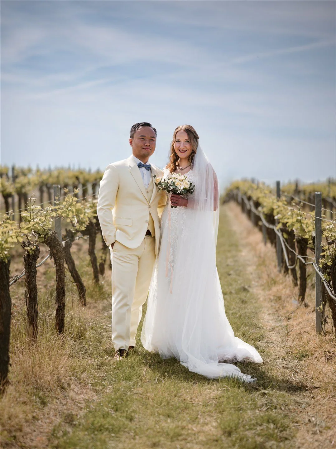 A couple in light coloured clothes stand between rows of vines.