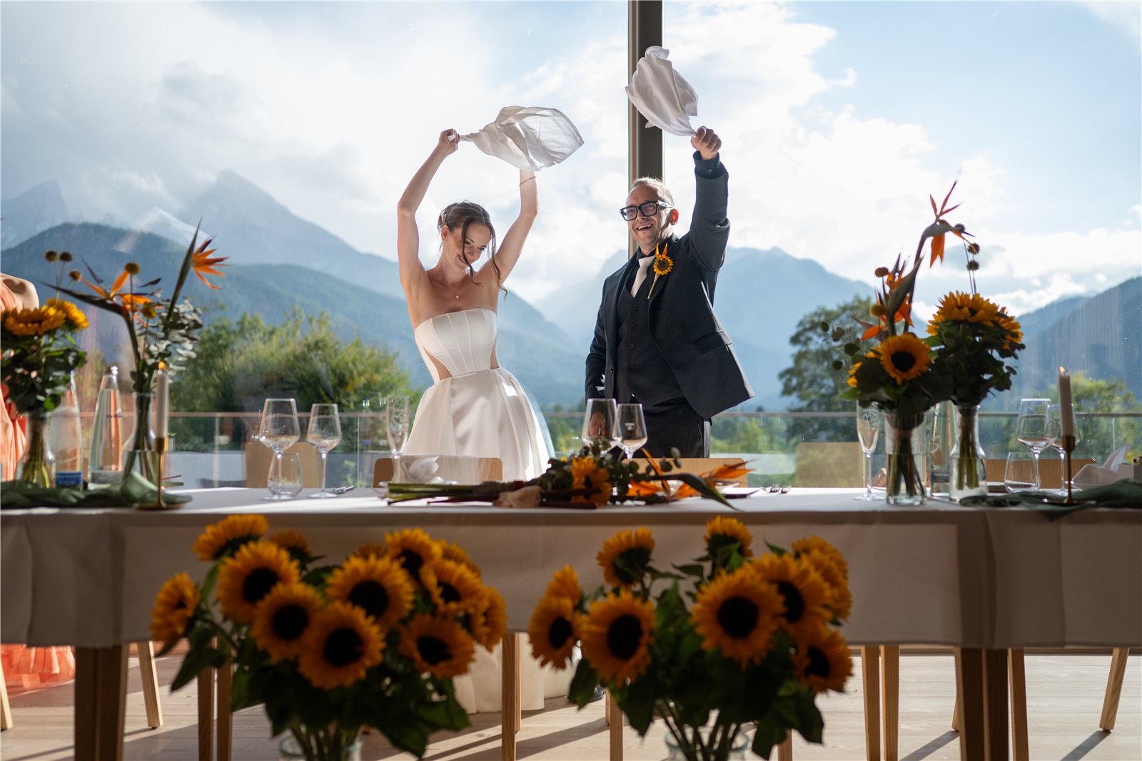 Ein Paar schwenkt Tücher an einer blumengeschmückten Tafel vor Bergblick.